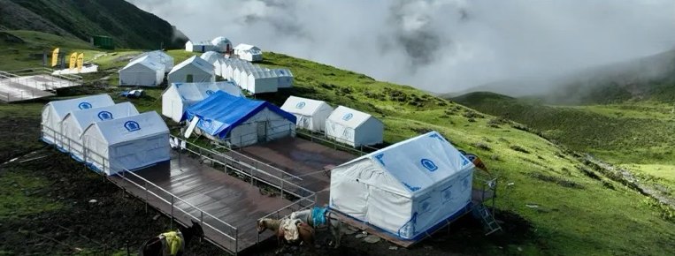 Vue aérienne du camp de base du Siguniang : des tentes blanches sur des plateformes en bois, sur une colline verte où monte la brume.