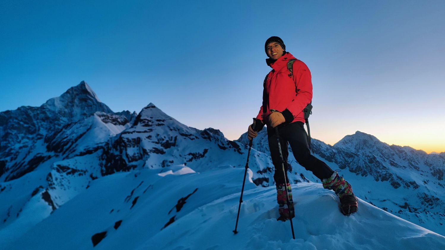 Un grimpeur équipé de crampons et de guêtres sourit sur une crête enneigée à l'aube, avec les sommets du Siguniang derrière lui.