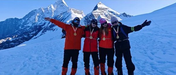 Quatre grimpeurs en vestes rouges, bras écartés sur un champ de neige, avec les sommets du Siguniang derrière eux.