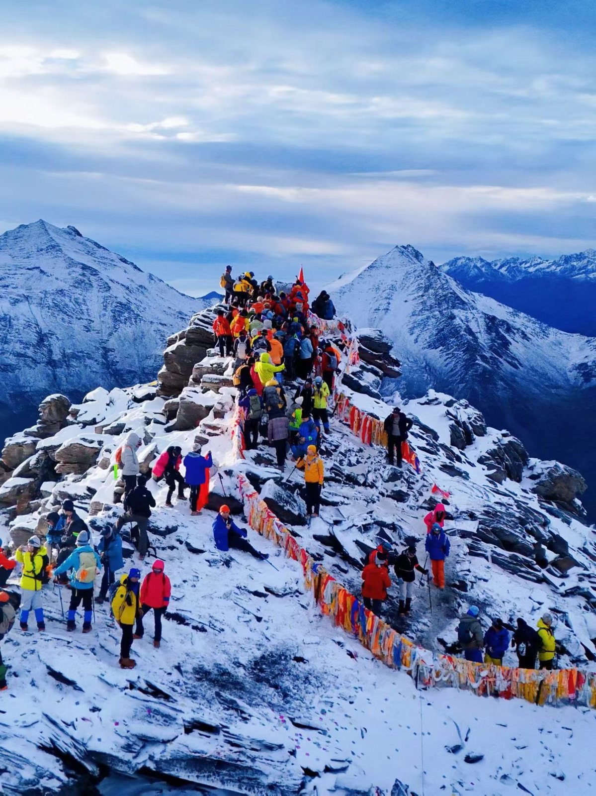 Des dizaines de grimpeurs en vestes colorées sur la crête sommitale enneigée de l'Erfeng, avec des drapeaux de prière, pendant les vacances nationales chinoises.