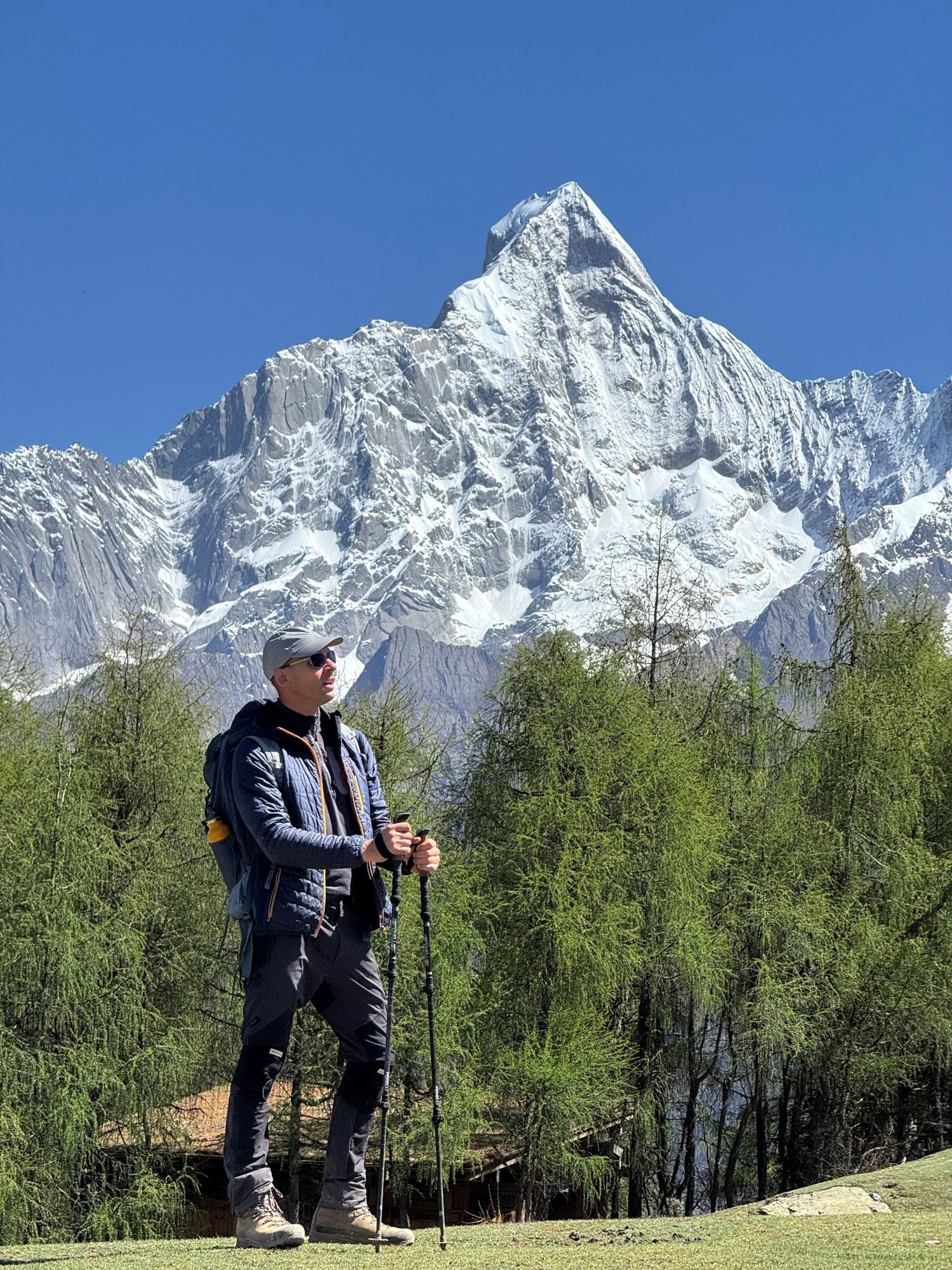 Un randonneur avec des bâtons de marche dans une prairie de mélèzes de la zone panoramique du Mont Siguniang, avec un sommet enneigé derrière lui.