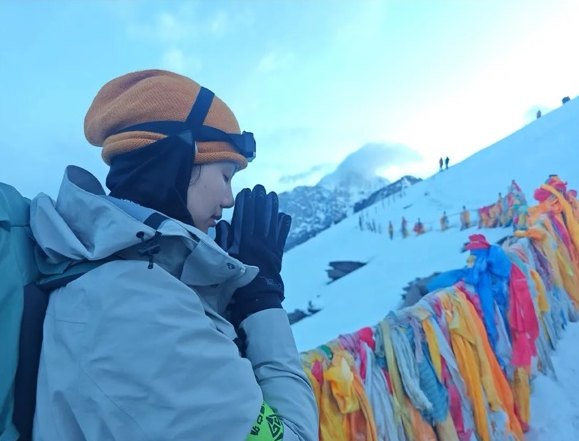 Un grimpeur en bonnet et lunettes de glacier fait une pause, les mains jointes, près de drapeaux de prière colorés sur une crête enneigée du Siguniang.