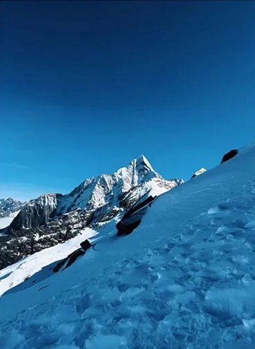 Un pic neigeux pointu du massif du Siguniang vu au-delà d'une pente de neige bleutée et raide, sous un ciel dégagé.