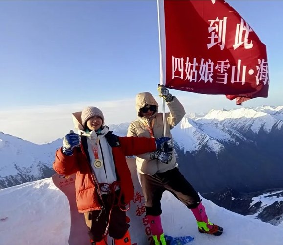 Deux grimpeurs tiennent un drapeau rouge au sommet du Siguniang, avec les chaînes enneigées du Sichuan derrière eux.